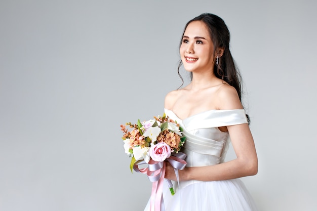 Jeune femme asiatique séduisante, bientôt mariée, vêtue d'une robe de mariée blanche à l'air heureux tenant un bouquet de fleurs. Concept pour la photographie avant le mariage.