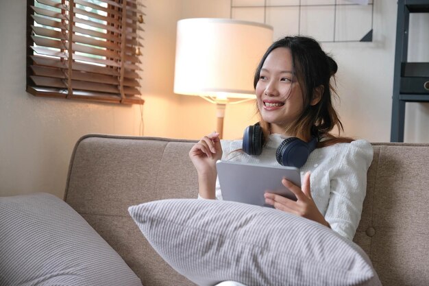 Une jeune femme asiatique séduisante avec un casque sans fil se détend dans le salon minimaliste à l'aide d'une tablette numérique