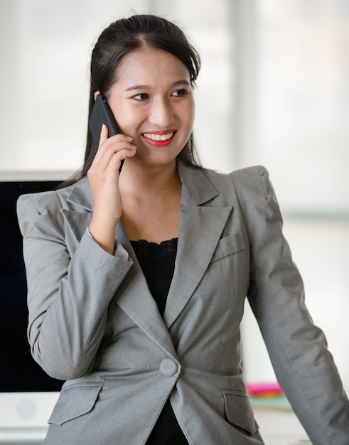 Jeune femme asiatique séduisante en costume d'affaires gris parlant au téléphone portable dans un bureau moderne avec un arrière-plan flou de fenêtres. Concept de style de vie de bureau moderne.