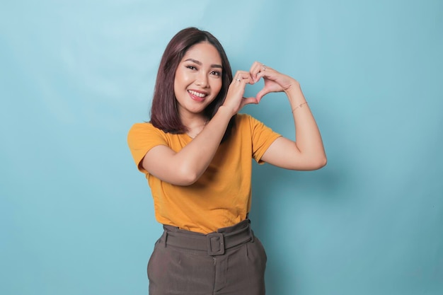 Une jeune femme asiatique séduisante se sent heureuse et romantique. Le geste du cœur exprime des sentiments tendres. Elle porte un t-shirt jaune décontracté sur fond bleu. Concept d'affection et de soins des personnes.