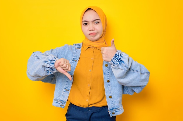 Jeune femme asiatique sérieuse en veste de jeans montrant les pouces vers le haut et vers le bas décision isolée sur fond jaune