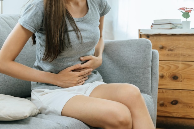 Photo jeune femme asiatique souffrant de maux d'estomac assis sur un canapé dans le salon à la maison personnes douloureuses maux d'estomac gynécologie douleurs menstruelles concept médical et de soins de santé