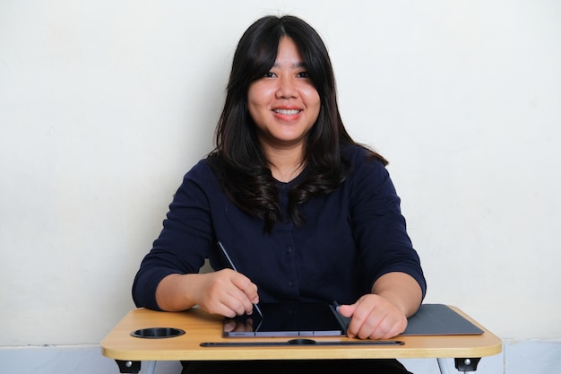 Photo jeune femme asiatique souriant à la caméra lorsqu'elle travaille à l'aide d'une tablette mobile