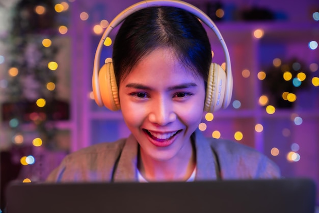 Une jeune femme asiatique souriante et heureuse écoute de la musique avec des écouteurs jaunes et des mains à l'aide d'un ordinateur portable en vacances dans la maison