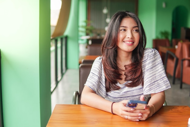 Une jeune femme asiatique souriante portant une robe et se sentant heureuse assise dans un restaurant pour un petit déjeuner tout en tenant un smartphone