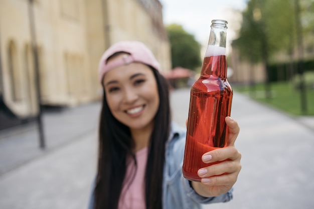 Jeune femme asiatique souriante tenant une bouteille de boisson rouge à l'extérieur