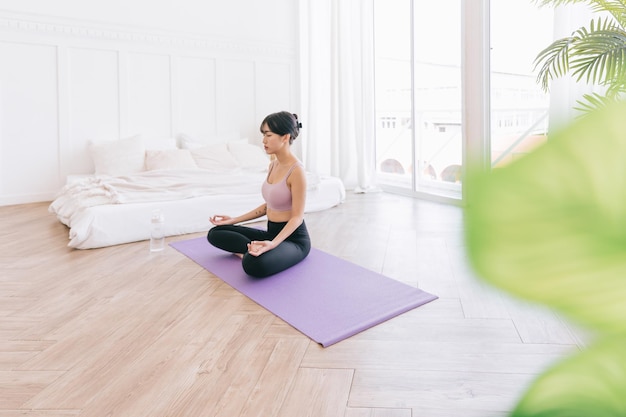 Jeune femme asiatique sportive séduisante et souriante pratiquant le yoga sur un tapis de yoga faisant de l'exercice Ardha Padmasana méditant dans la pose de demi-lotus à l'intérieur à la maison portant des vêtements de sport