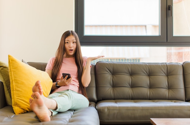 Jeune femme asiatique à la surprise et choqué, avec la mâchoire tombée tenant un objet avec une main ouverte sur le côté