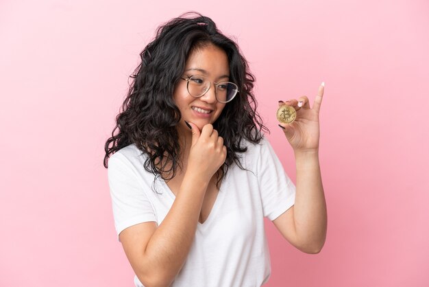 Jeune femme asiatique tenant un Bitcoin isolé sur fond rose regardant sur le côté et souriant