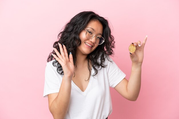 Jeune femme asiatique tenant un Bitcoin isolé sur fond rose saluant avec la main avec une expression heureuse