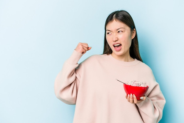 Jeune femme asiatique tenant un bol de céréales isolé sur fond bleu levant le poing après un concept de vainqueur de la victoire