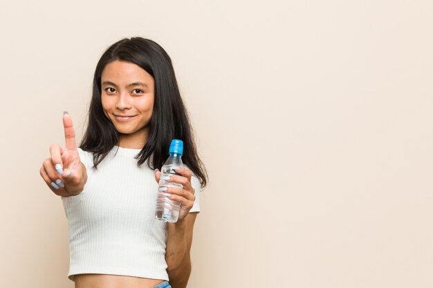 Jeune femme asiatique tenant une bouteille d'eau montrant le numéro un avec le doigt.