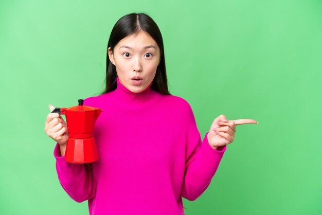 Jeune femme asiatique tenant une cafetière sur fond isolé chroma key surpris et pointant vers le côté