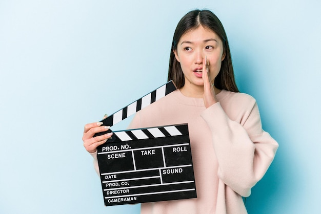 Jeune femme asiatique tenant un clap isolé sur fond bleu dit une nouvelle secrète sur le freinage à chaud et regarde de côté