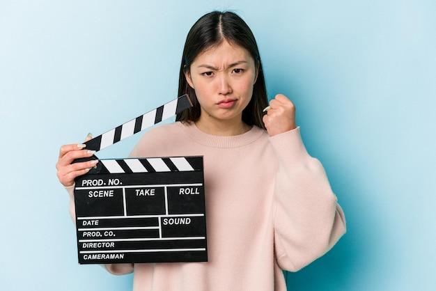 Jeune femme asiatique tenant un clap isolé sur fond bleu montrant le poing à la caméra expression faciale agressive