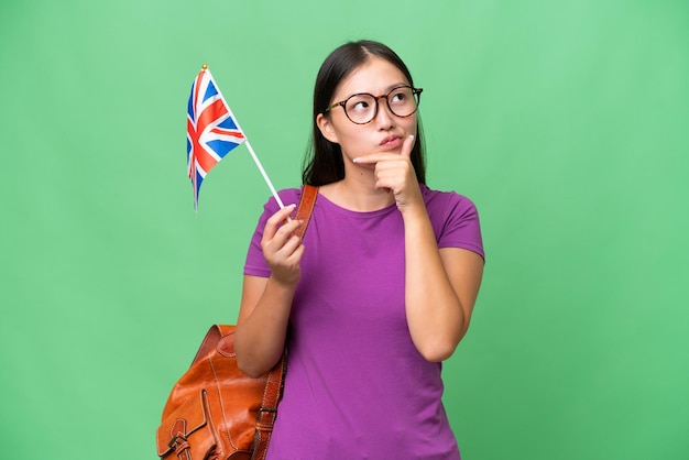 Jeune femme asiatique tenant un drapeau du Royaume-Uni sur fond isolé ayant des doutes
