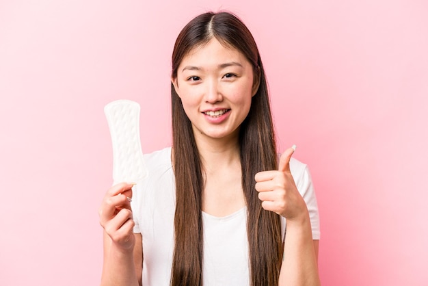 Jeune femme asiatique tenant une serviette hygiénique isolée sur fond rose souriant et levant le pouce vers le haut