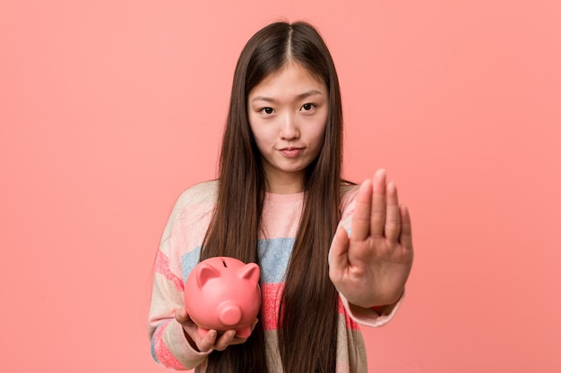 Jeune femme asiatique tenant une tirelire debout avec la main tendue montrant le panneau d'arrêt, vous empêchant.