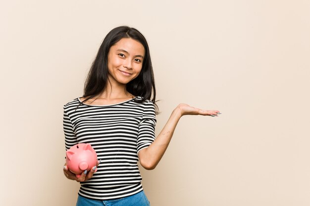 Jeune femme asiatique tenant une tirelire montrant un espace de copie sur une paume et tenant une autre main sur la taille.