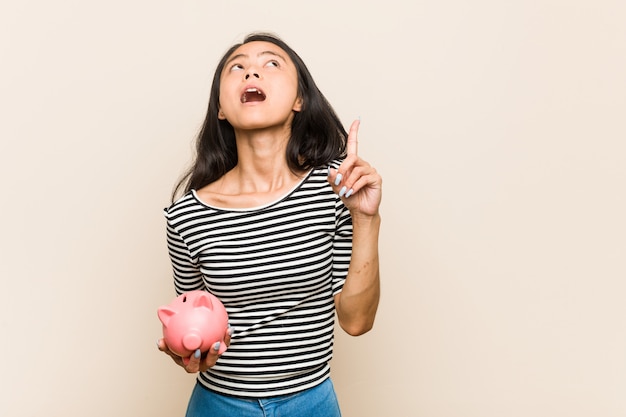 Jeune femme asiatique tenant une tirelire pointant à l'envers avec la bouche ouverte.