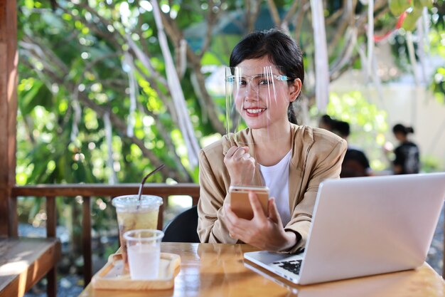 Jeune femme asiatique en tenue décontractée avec écran facial pour les soins de santé, assise dans un café et utilisant un smartphone et travaillant sur un ordinateur portable. Nouveau concept normal et de distanciation sociale