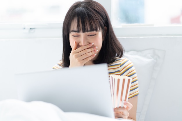 Jeune femme asiatique utilisant un ordinateur portable pour regarder des émissions de télévision, manger du pop-corn à la maison