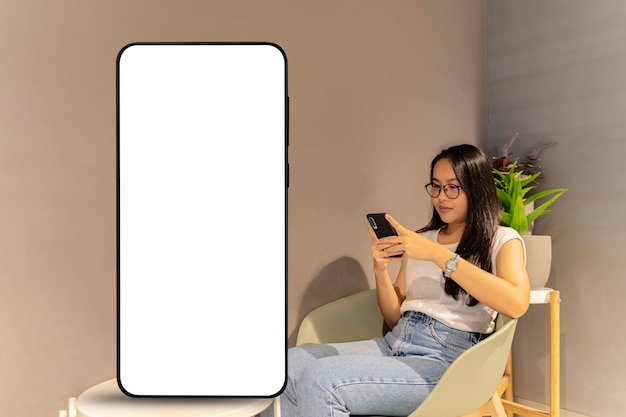 Jeune femme asiatique utilisant un smartphone se relaxant dans un fauteuil près d'un téléphone portable géant avec une maquette d'écran vide Technologie en ligne et concept de style de vie mobile