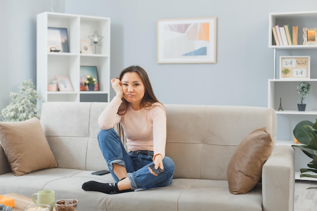 Jeune femme asiatique en vêtements décontractés assis sur un canapé à l'intérieur de la maison en train de manger des biscuits tenant à distance en regardant la télévision l'air fatigué et ennuyé