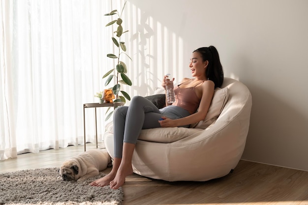 Une jeune femme asiatique en vêtements de sport buvant de l'eau pure dans un verre dans le salon à la maison.