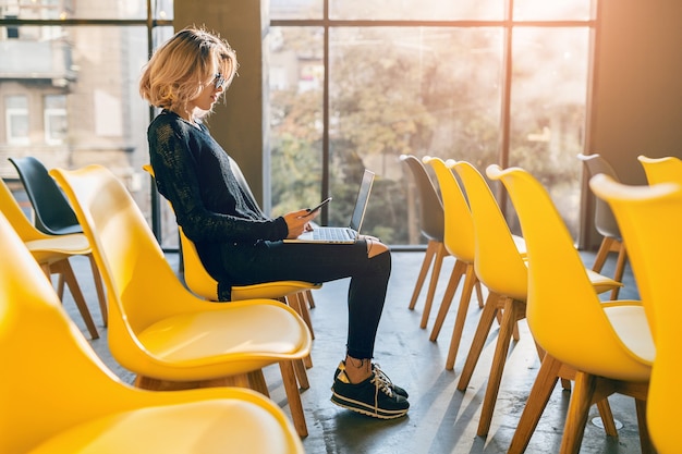Jeune femme assez occupée assise seule dans la salle de conférence, de nombreuses chaises jaunes, travaillant sur un ordinateur portable