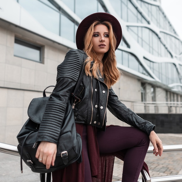 Jeune femme assez séduisante en veste de cuir noir en pantalon avec un sac à dos élégant dans un élégant chapeau posant