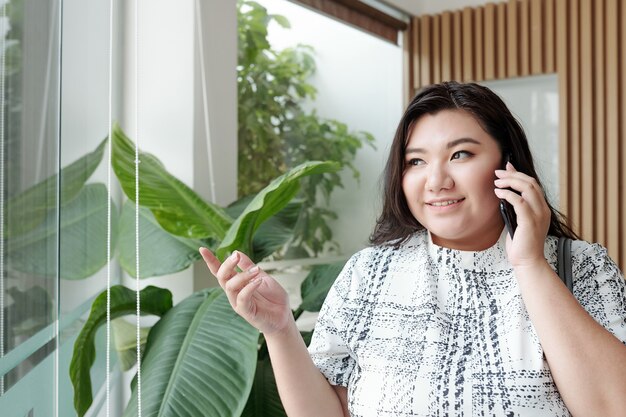 Jeune femme assez souriante parlant au téléphone avec un ami d'un collègue et regardant par la fenêtre d'un hôtel ou d'un immeuble de bureaux