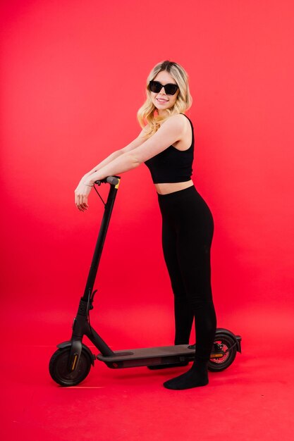 Jeune femme assez souriante en tenue de sport debout avec un scooter électrique regardant joyeusement à huis clos