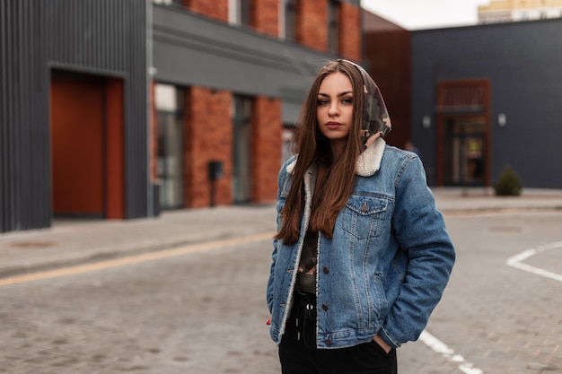 Jeune femme assez urbaine à la mode en veste en jean bleu jeunesse pose près du bâtiment de la ville. Belle fille américaine en jeans élégants dans la rue. Vêtements de mode décontractés pour femmes.