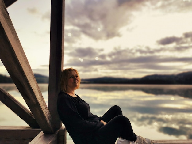 Une jeune femme assise au bord de la mer contre le ciel au coucher du soleil
