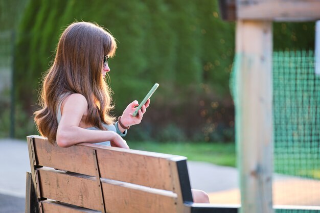 Jeune femme assise sur un banc de parc parcourant son téléphone portable à l'extérieur par une chaude soirée d'été. Concept de communication et de connexion mobile.