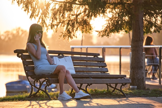 Jeune femme assise sur un banc de parc parlant sur son téléphone portable à l'extérieur par une chaude soirée d'été. Concept de communication et de connexion mobile.