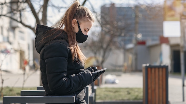 Jeune femme assise sur un banc de rue avec masque médical et vêtements noirs