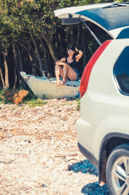 Jeune femme assise sur un bateau à la plage près de la voiture