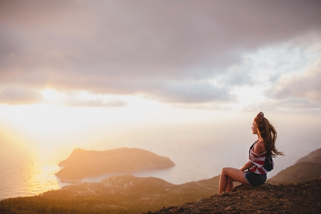 Jeune femme assise sur le bord de la falaise, regardant le coucher du soleil sur la mer Méditerranée