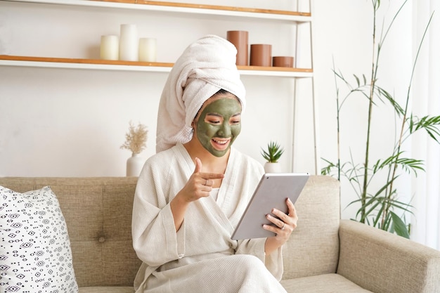 Jeune femme assise sur le canapé dans le salon, elle utilise un masque facial et profite de sa journée