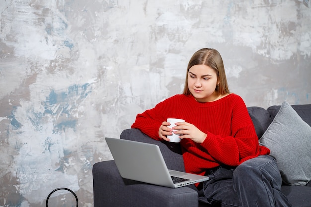 Jeune femme assise sur le canapé en regardant l'écran du portable. Une étudiante indépendante motivée et à domicile travaillant sur un ordinateur en ligne.