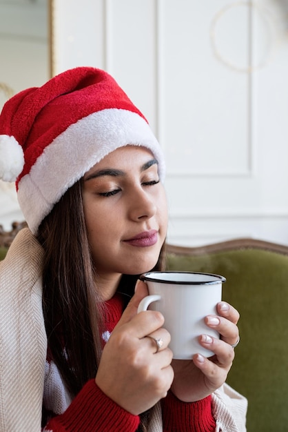 Jeune femme assise sur un canapé seule dans un salon décoré pour Noël ayant une tasse de boisson chaude