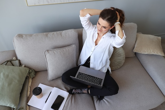 Jeune femme assise sur le canapé et travaillant sur un ordinateur portable à la maison