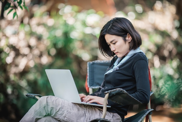 Jeune femme assise sur une chaise de camping et utilisant un ordinateur portable tout en vous relaxant sur le camping dans l'espace de copie de la forêt