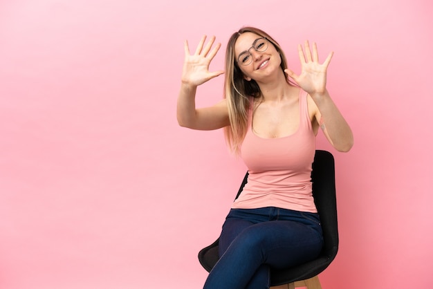 Jeune femme assise sur une chaise sur fond rose isolé comptant dix avec les doigts