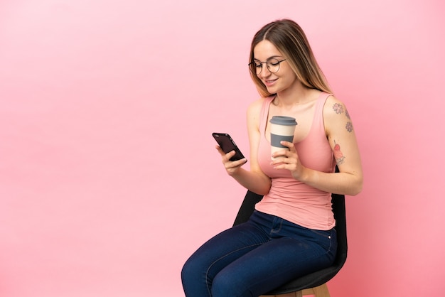 Jeune femme assise sur une chaise sur fond rose isolé tenant du café à emporter et un mobile