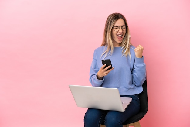 Jeune femme assise sur une chaise avec ordinateur portable sur fond rose isolé avec téléphone en position de victoire