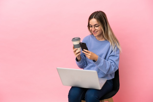 Jeune Femme Assise Sur Une Chaise Avec Ordinateur Portable Sur Fond Rose Isolé Tenant Du Café à Emporter Et Un Mobile