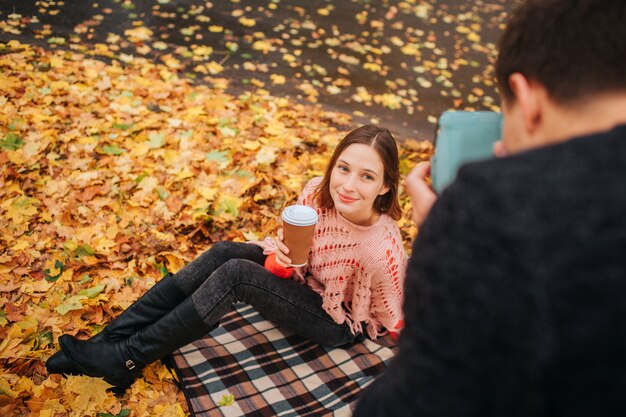 Jeune femme assise sur une couverture au sol et pose devant la caméra. Elle tient une tasse de café à la main. Un jeune homme en vêtements noirs prend des photos d'elle.